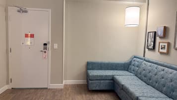 A room with a light-colored door, a blue sectional sofa, a floor lamp, and framed pictures on the wall.