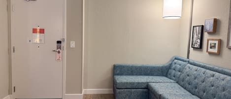 A room with a light-colored door, a blue sectional sofa, a floor lamp, and framed pictures on the wall.
