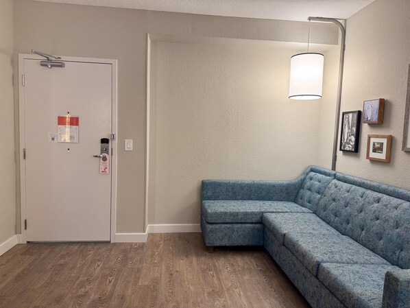 Una habitación con una puerta de color claro, un sofá seccional azul, una lámpara de pie y cuadros en la pared.