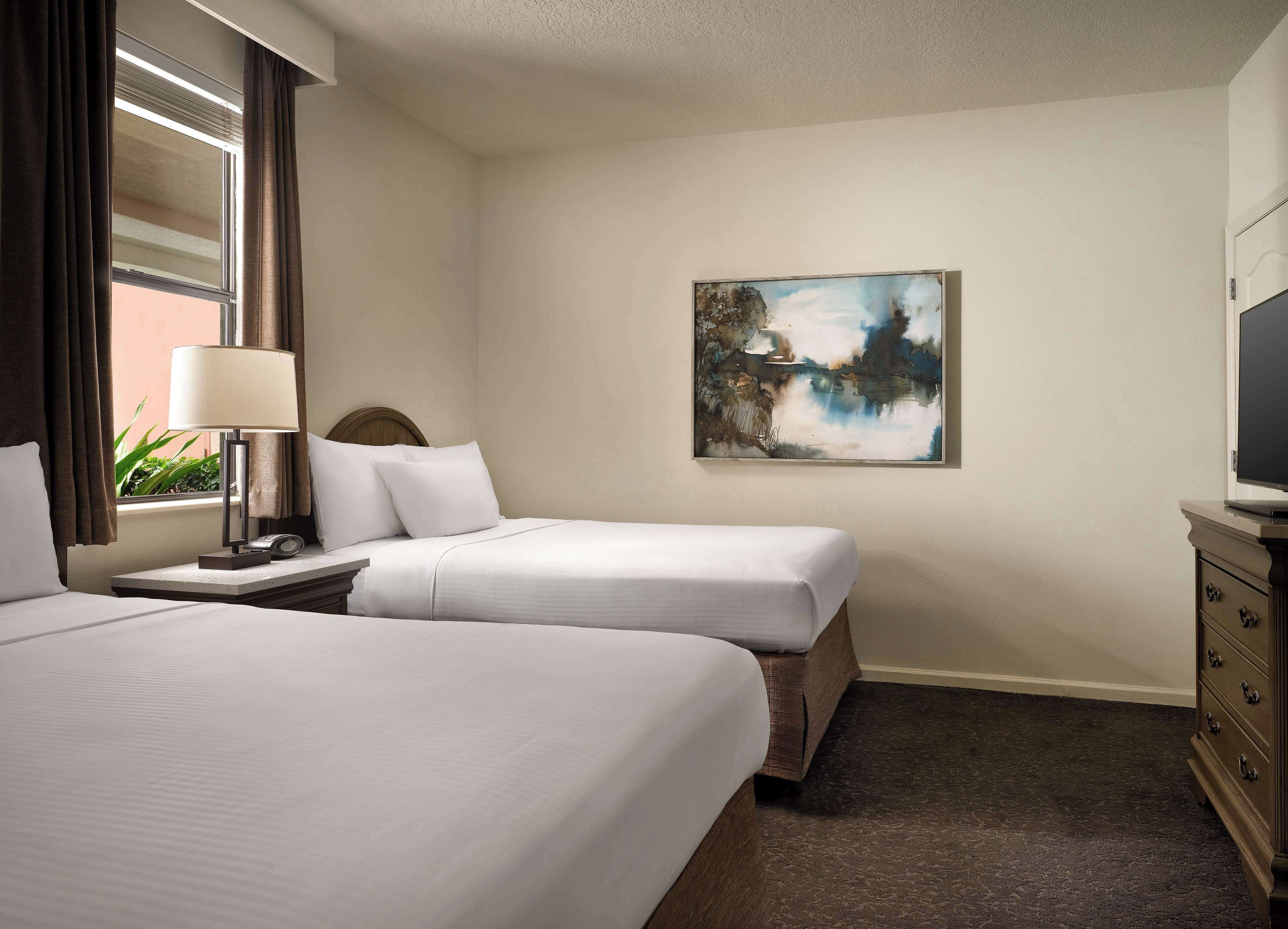 A hotel room with two beds, a nightstand, a lamp, a painting on the wall, and a dresser.