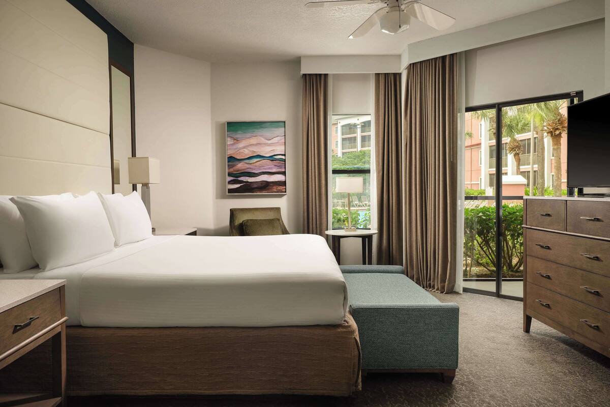 A hotel room with a bed, a desk, a chair, a dresser, a television, a painting, and a window with curtains.