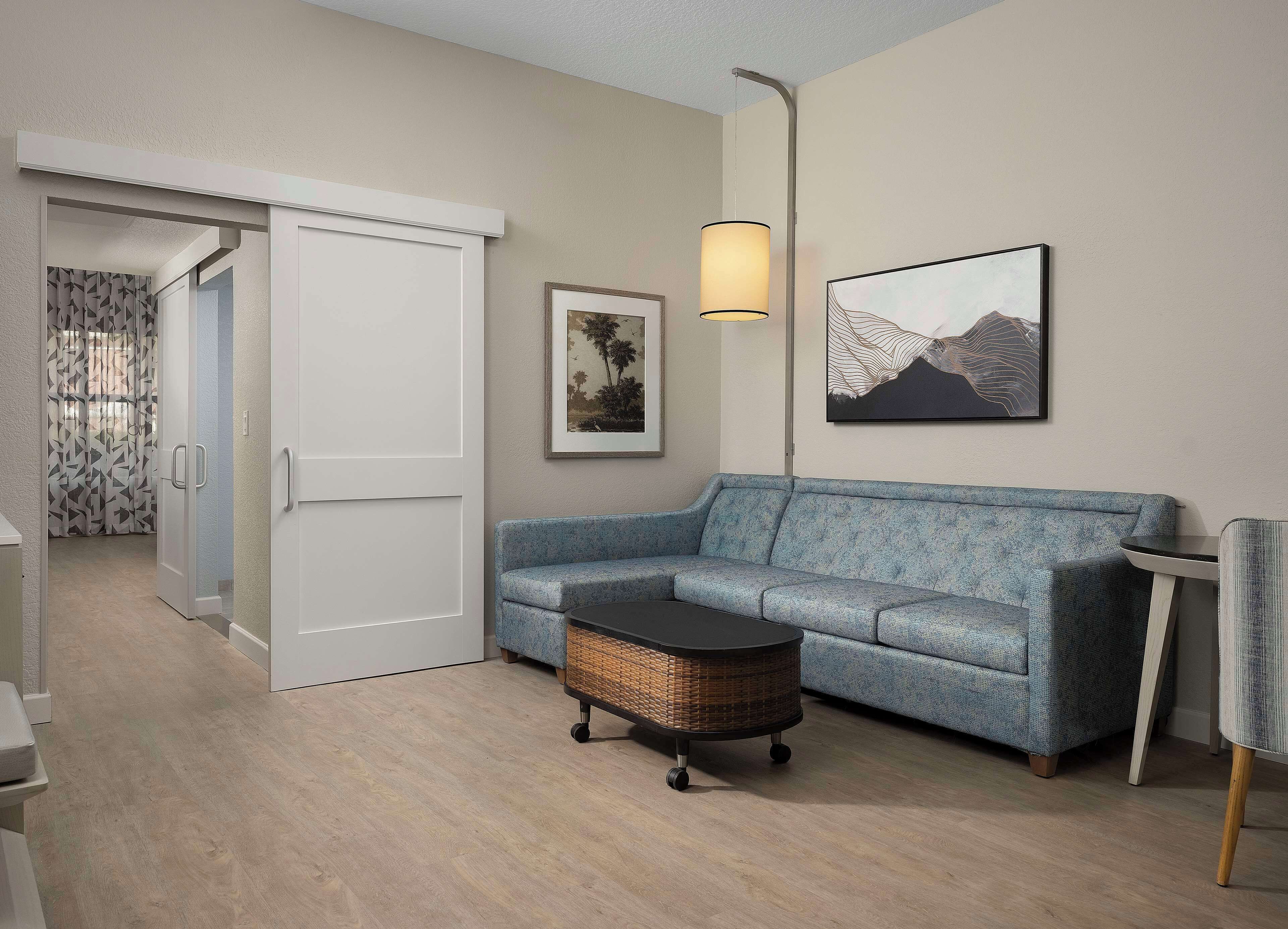 A room with a blue sofa, a wicker side table, a framed picture, a lamp, and a door leading to another room.