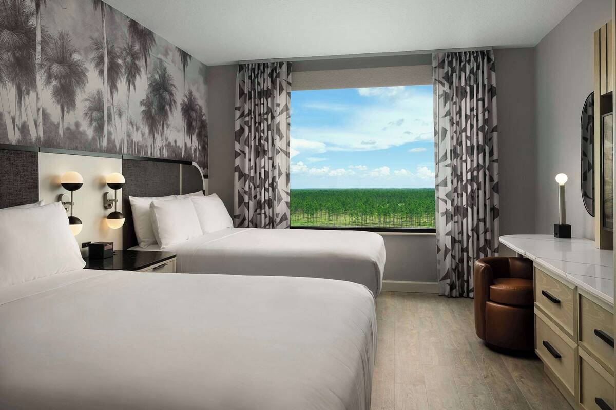 A hotel room with two beds, a desk, a chair, a nightstand, a lamp, and a view of a green landscape through the window.
