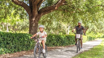 Duas pessoas andam de bicicleta por um caminho pavimentado, cercado por vegetação.