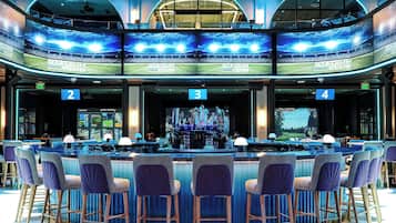 Zona de bar con diversas opciones de asientos, una barra central y una pantalla grande que muestra una vista de estadio.