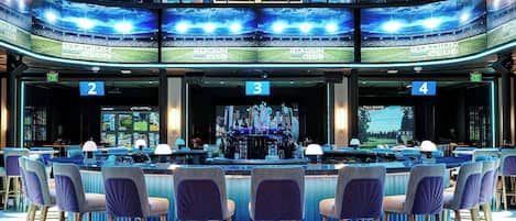 Zona de bar con diversas opciones de asientos, una barra central y una pantalla grande que muestra una vista de estadio.