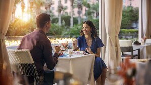 A couple dining at an outdoor table with candles, surrounded by greenery and a building in the background.