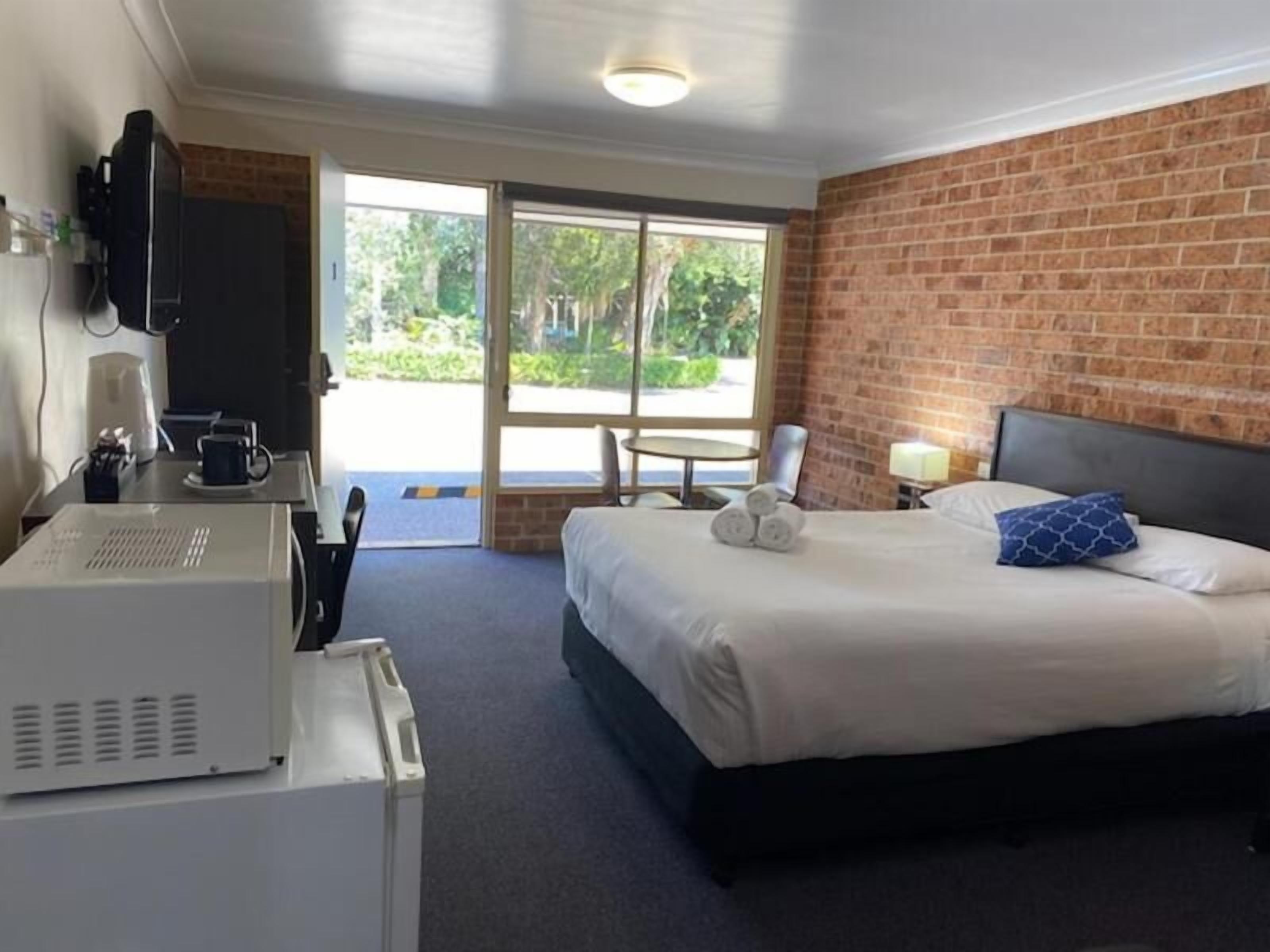 A hotel room with a bed, a desk with a computer, a television, and a window with a view of greenery.