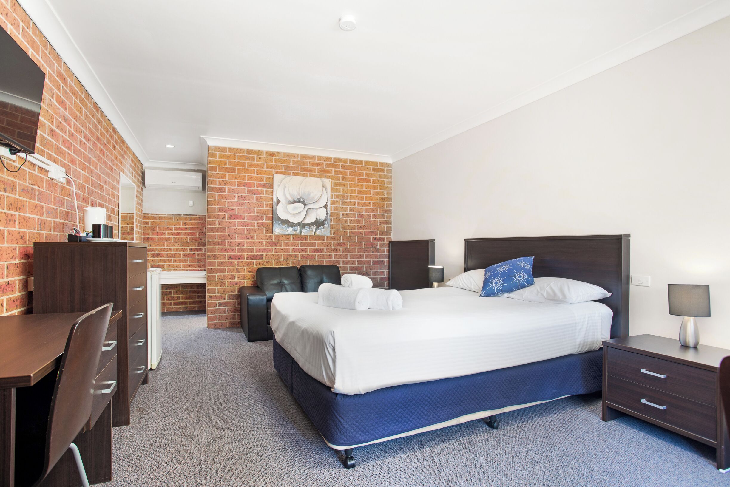 A hotel room with a brick wall, a bed with white linens, a desk with a chair, and a small table with a lamp.