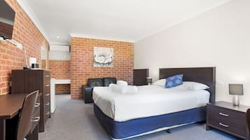 A hotel room with a brick wall, a bed with white linens, a desk with a chair, and a small table with a lamp.
