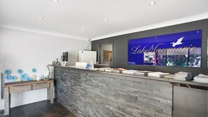 A reception area with a stone wall, a wooden desk, and a large screen displaying 'Lake Macquarie'. - Lake Macquarie Motor Inn (Marks Point)