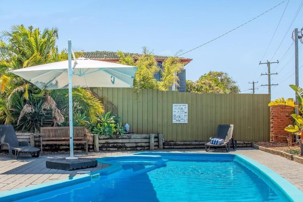 Une piscine entourée d’une clôture en bois et de verdure, avec des chaises longues et un parasol.