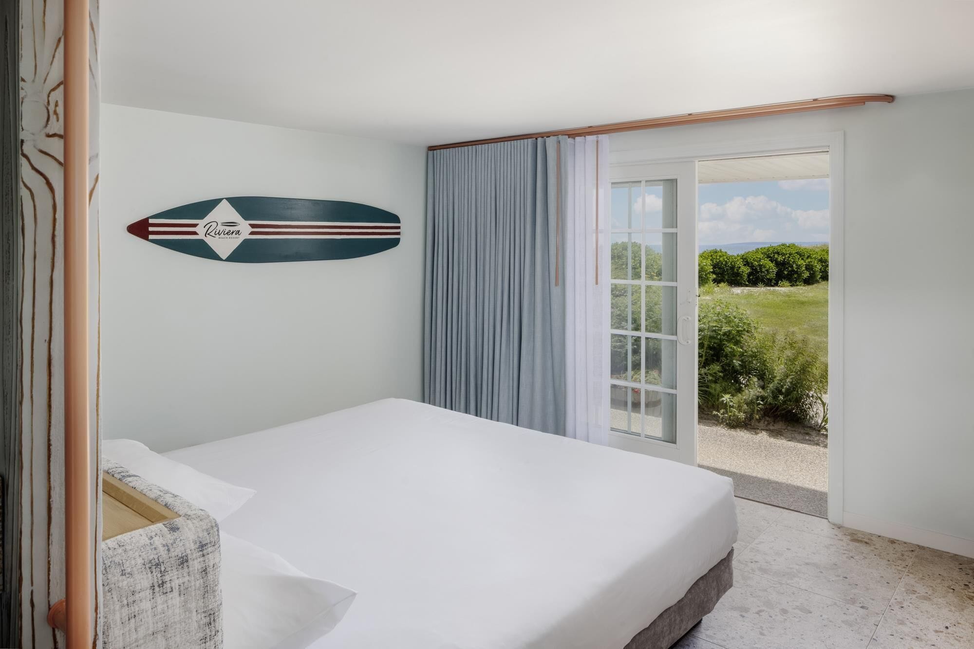 A bedroom with a bed, a view of the outdoors, and a surfboard decoration on the wall.