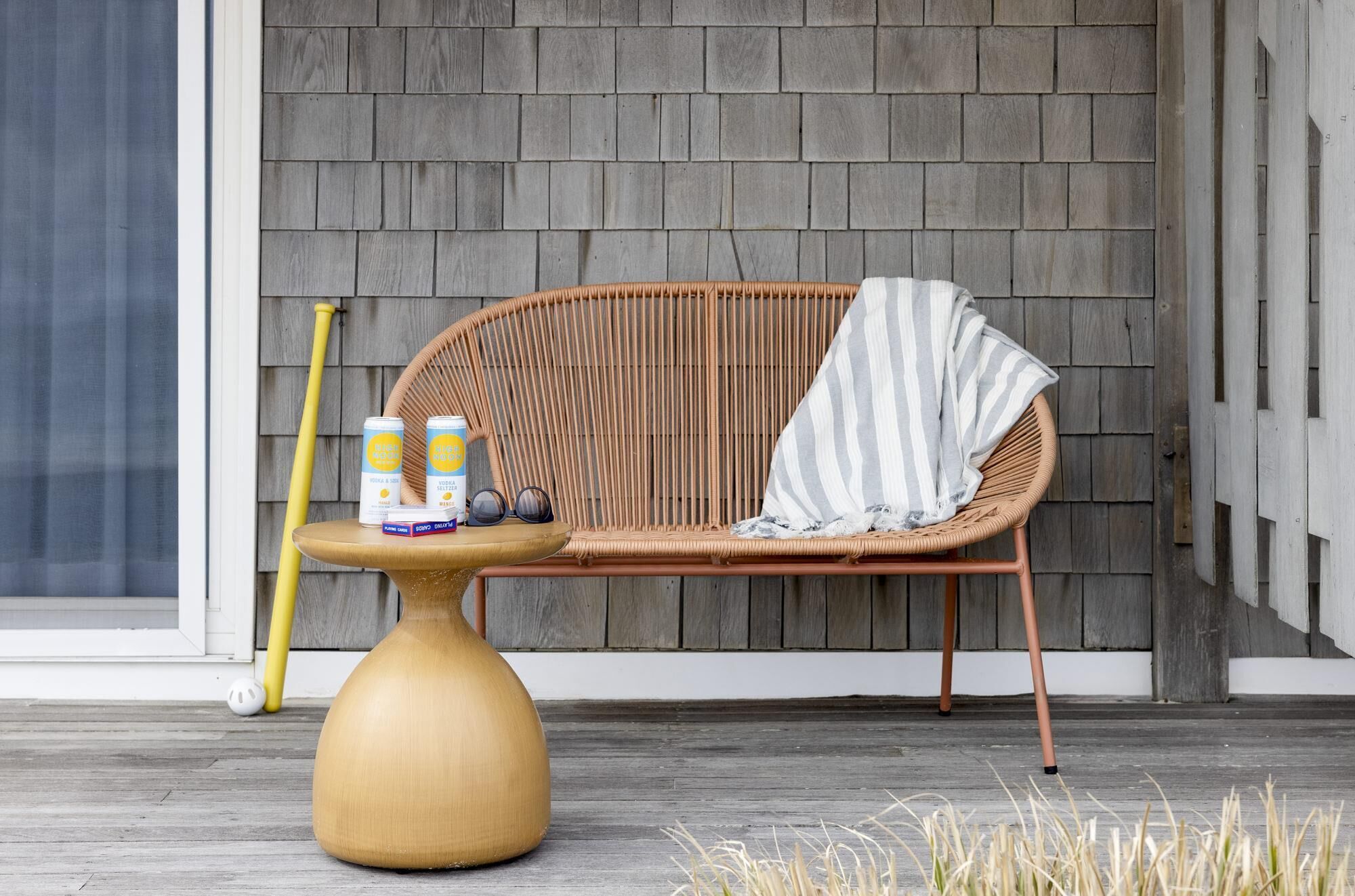 Une chaise en osier avec un coussin à rayures, une petite table ronde en bois sur laquelle est posé un vase jaune, une canette de soda et une paire de lunettes de soleil.