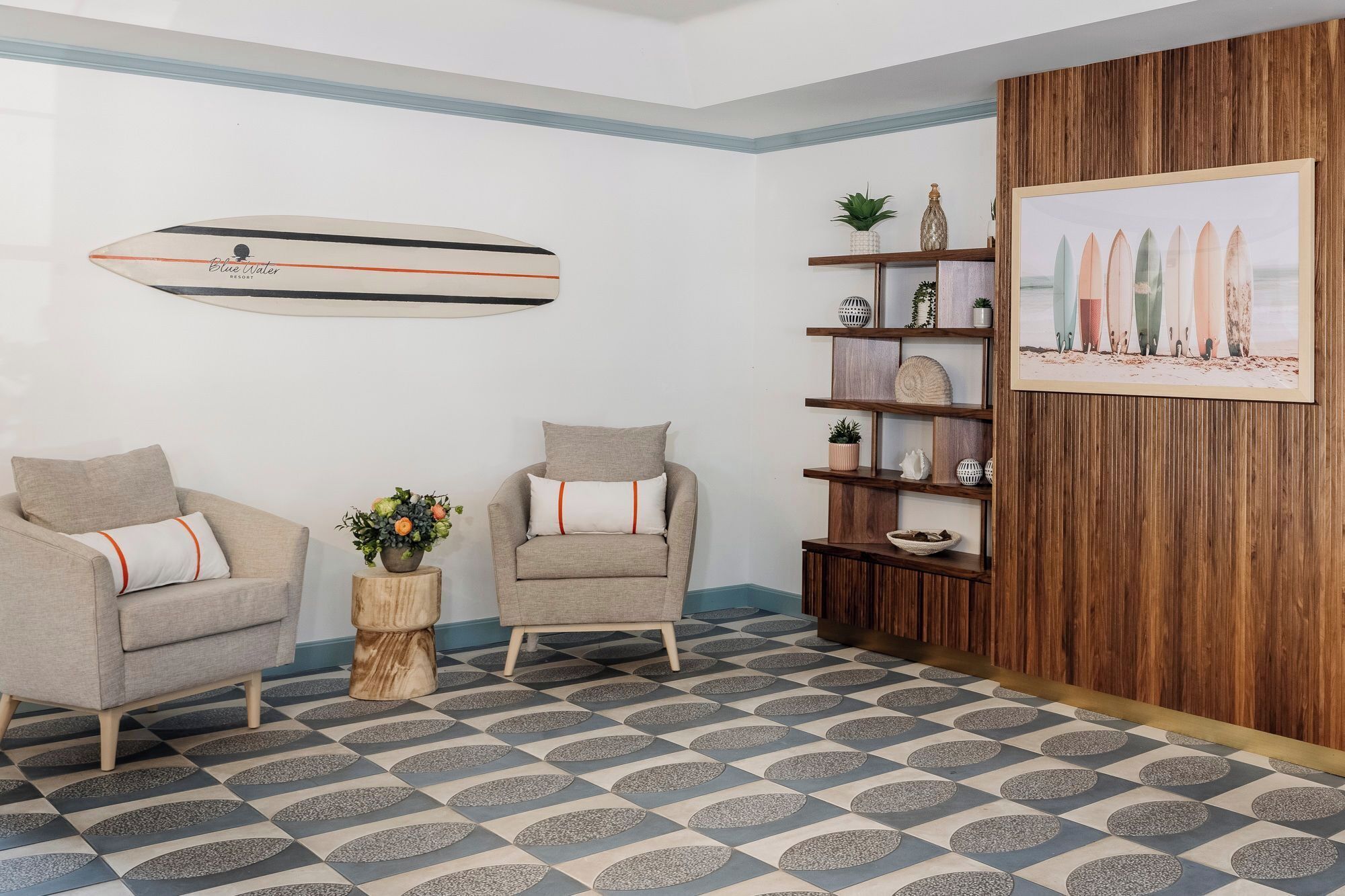 A living room with two armchairs, a coffee table, a surfboard, a wooden shelf with decorative items, and a framed picture of surfboards.