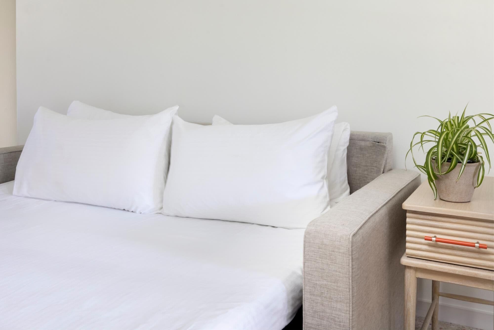 Un lit avec du linge de lit blanc, un canapé gris et une petite table de chevet en bois sur laquelle est posée une plante en pot.