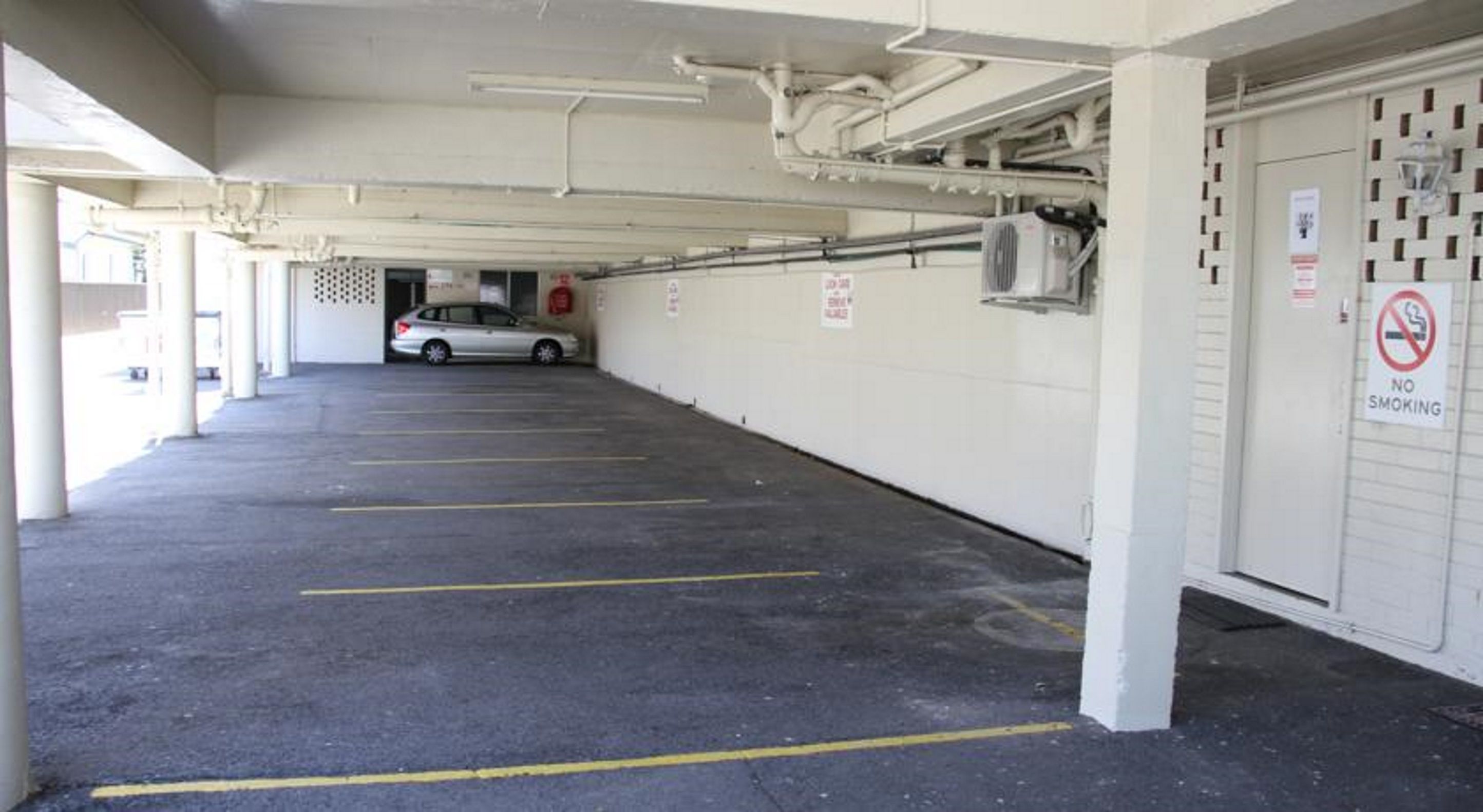 A parking garage with multiple parking spaces, a car in the distance, and a 'No Smoking' sign on the wall.