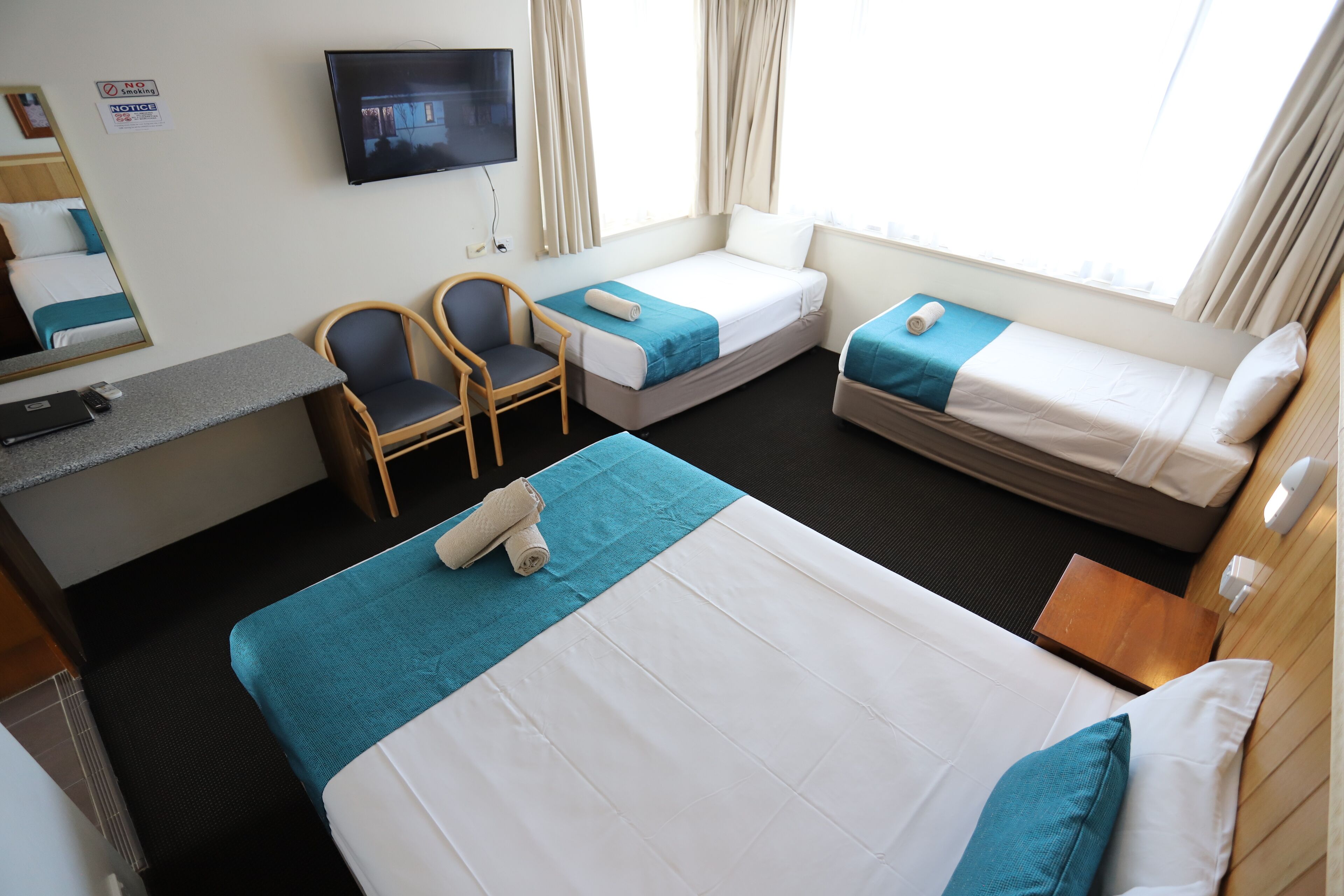 A hotel room with a large bed, two smaller beds, a desk with a chair, a TV, and a window with curtains.