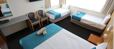 A hotel room with a large bed, two smaller beds, a desk with a chair, a TV, and a window with curtains.