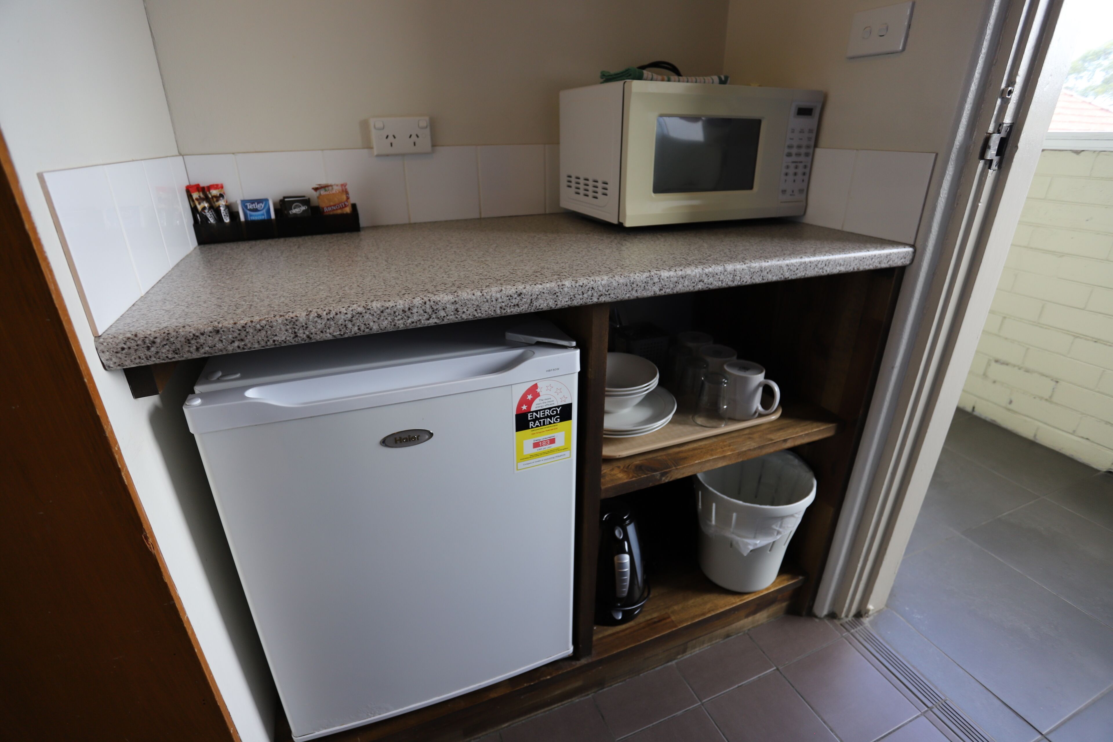 A compact kitchenette with a microwave, a small refrigerator, and a shelf with dishes and a kettle.