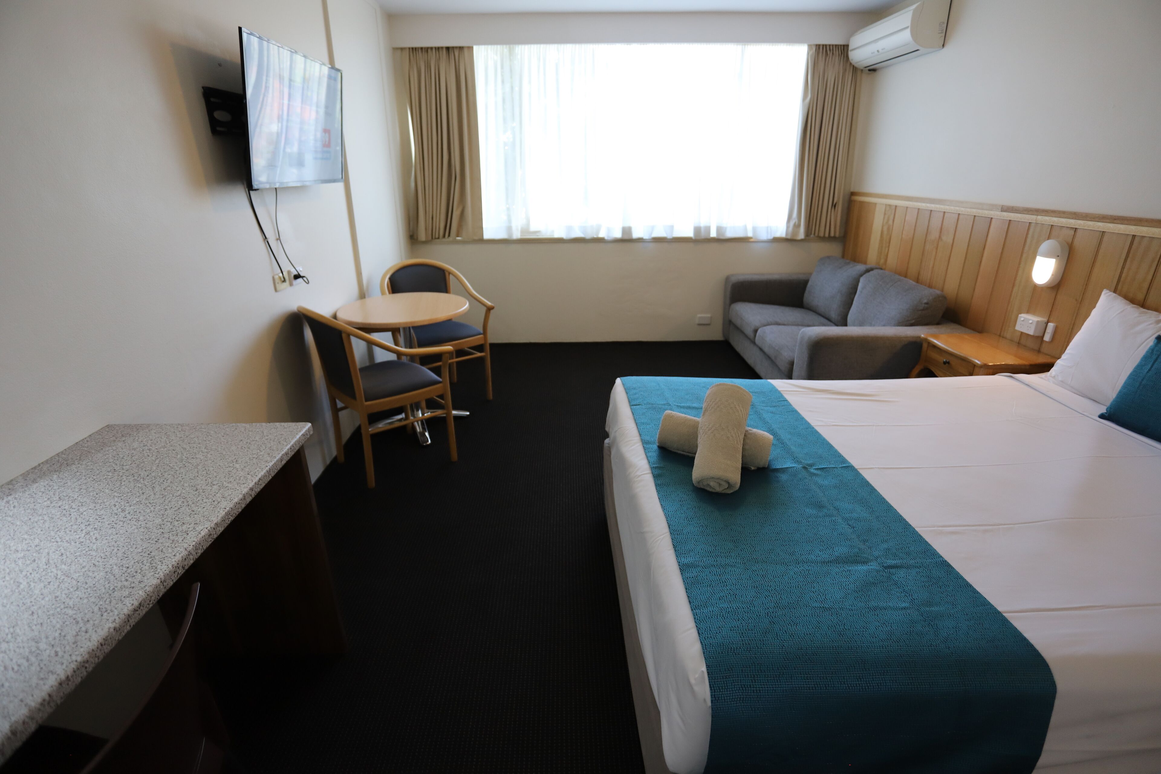 A hotel room with a bed, a desk, a chair, a sofa, and a TV mounted on the wall.