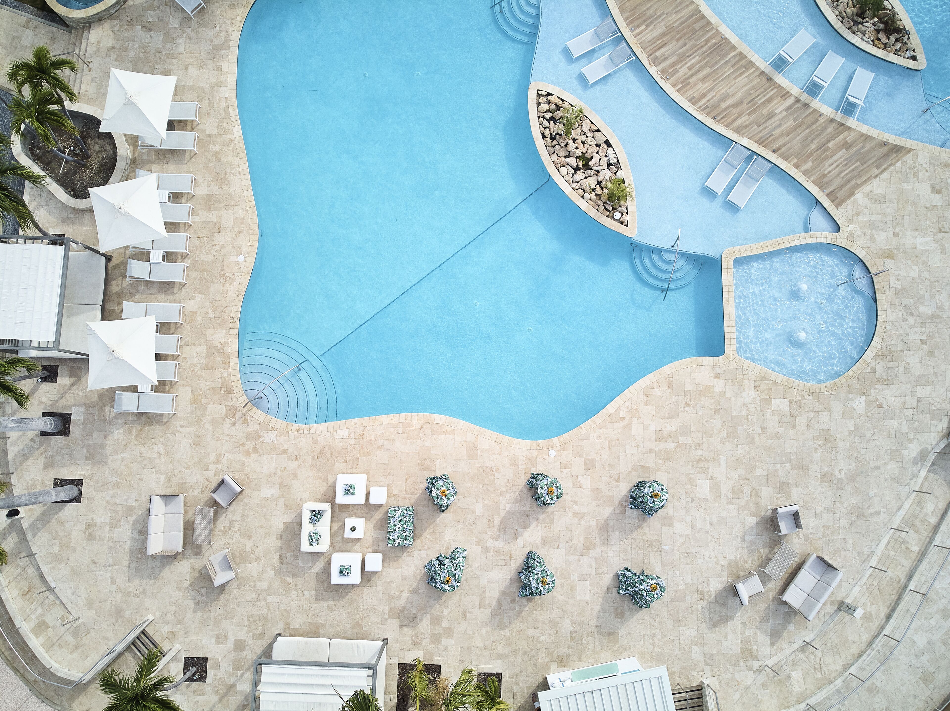 Vue aérienne d’un espace piscine d’un complexe hôtelier, avec différents coins aménagés pour s’asseoir, une petite fontaine et des palmiers tout autour.