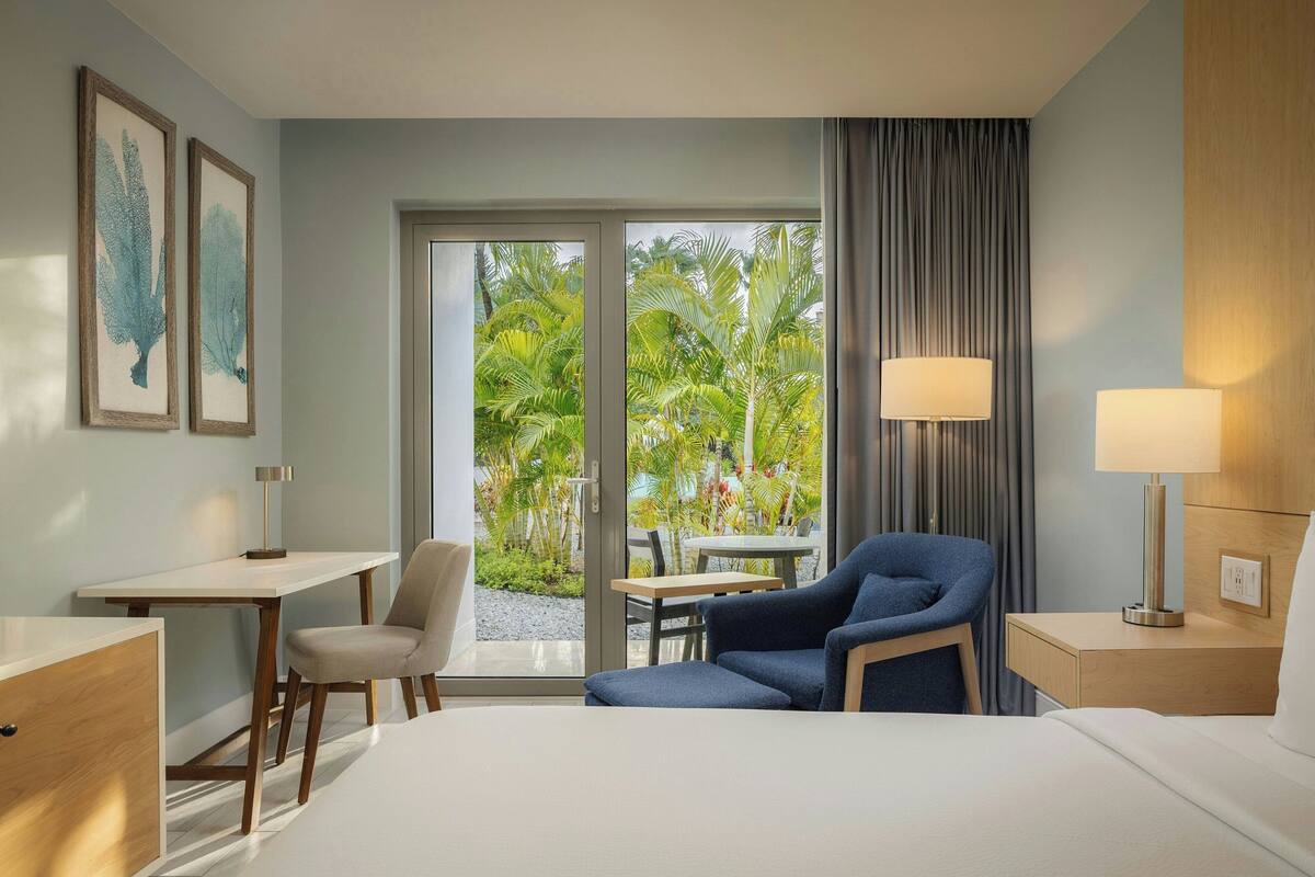 Habitación de hotel con cama, escritorio, silla, mesa y vista a palmeras a través de la ventana.