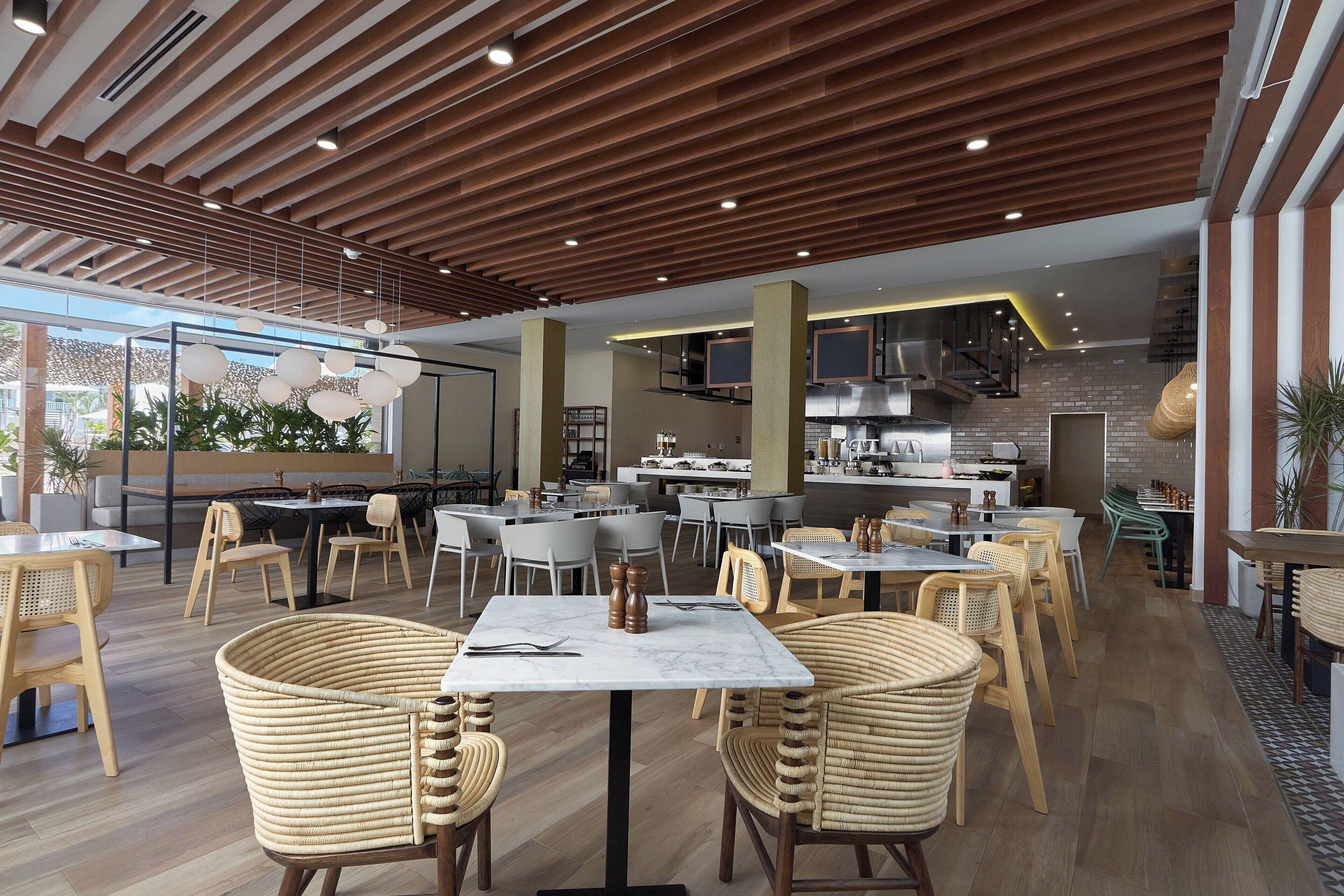 Un restaurante espacioso con vigas de madera en el techo, varias zonas de comedor con sillas de mimbre y una cocina a la vista.