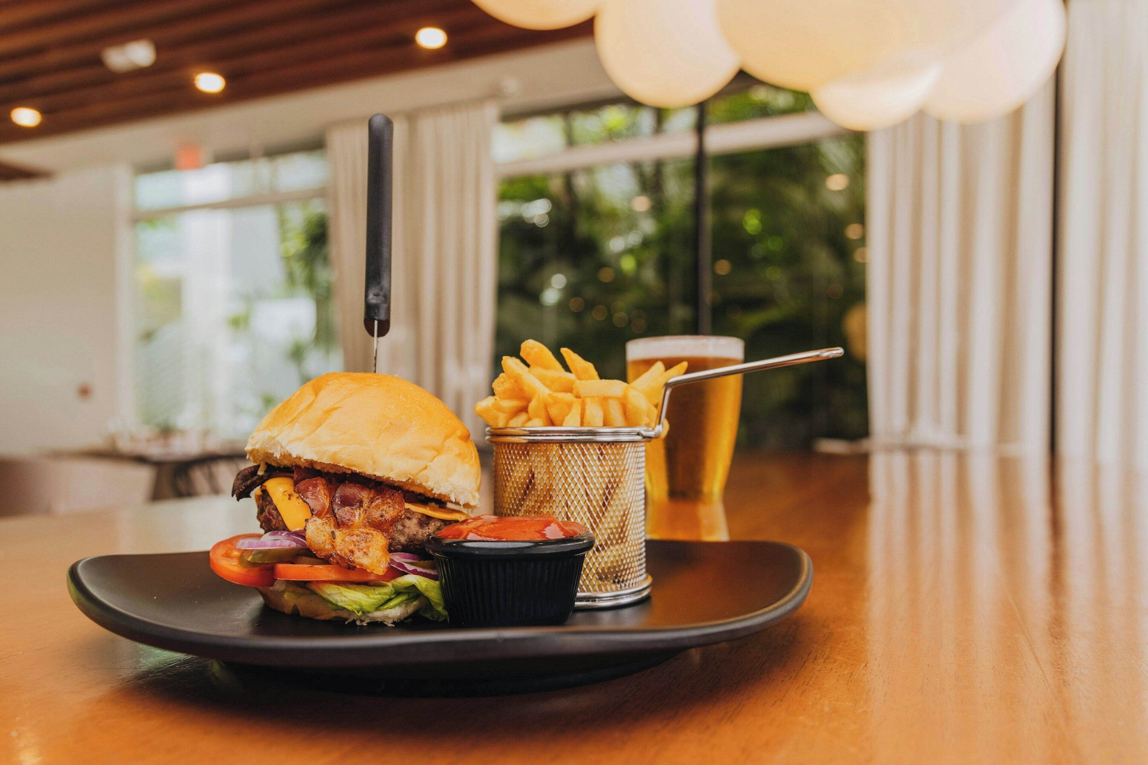 A meal consisting of a burger, fries, and a beer on a plate, with a window and curtains in the background.