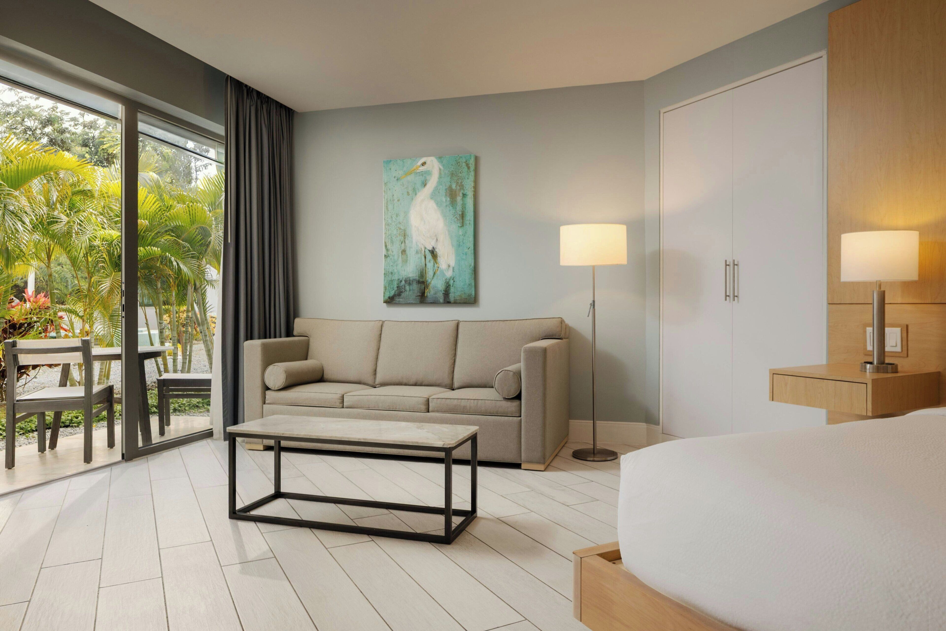 Une chambre d’hôtel avec un canapé, une table basse, un lit, un bureau, une lampe, un tableau et une vue sur la verdure.