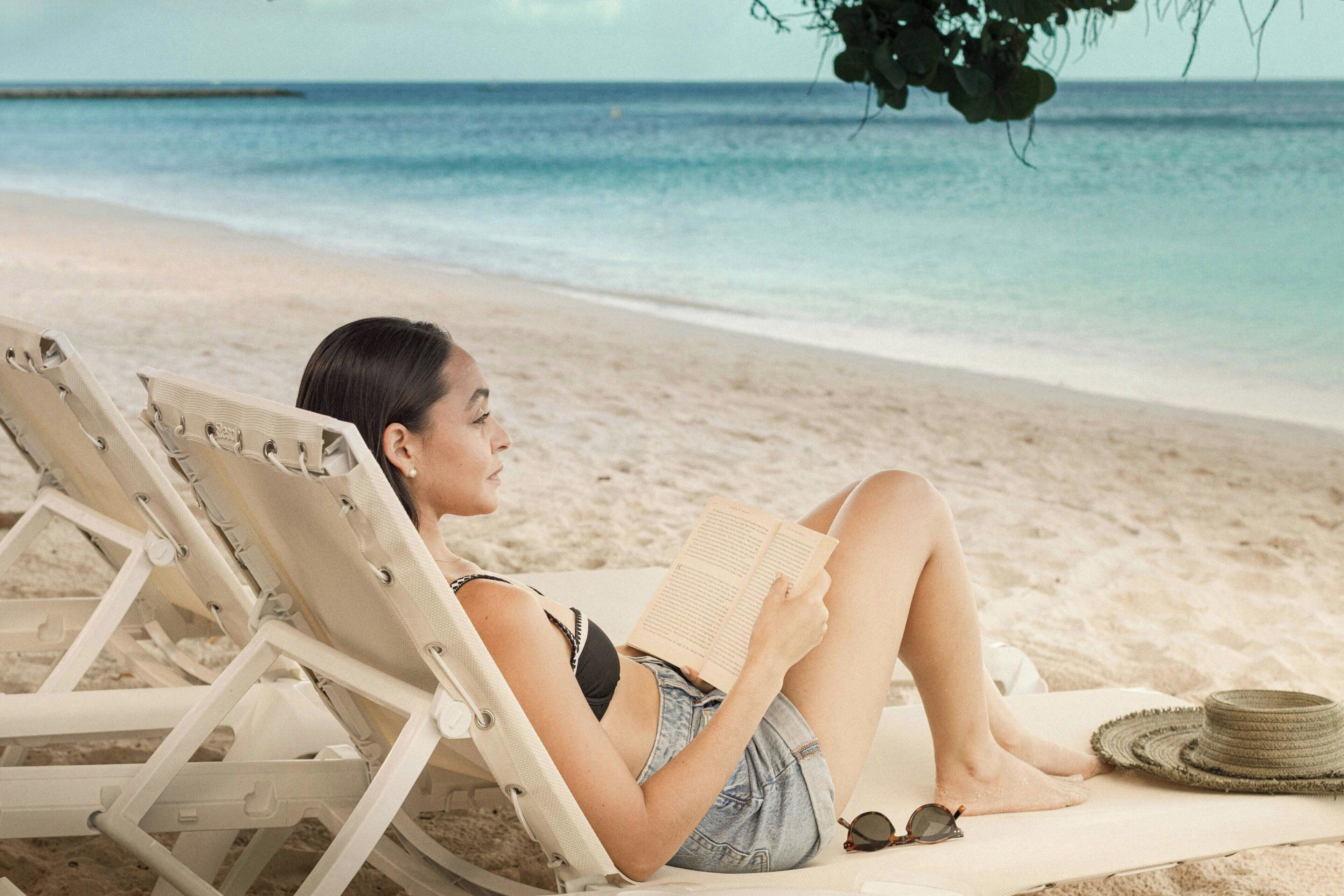 A person reclining on a beach chair, reading a book, with a straw hat and sunglasses beside them.