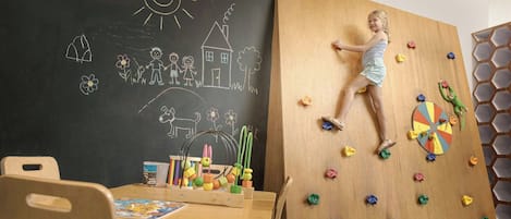 Un niño trepando una pared de escalada de madera con puntos de colores, una pizarra con dibujos y una mesa con juguetes y un libro.