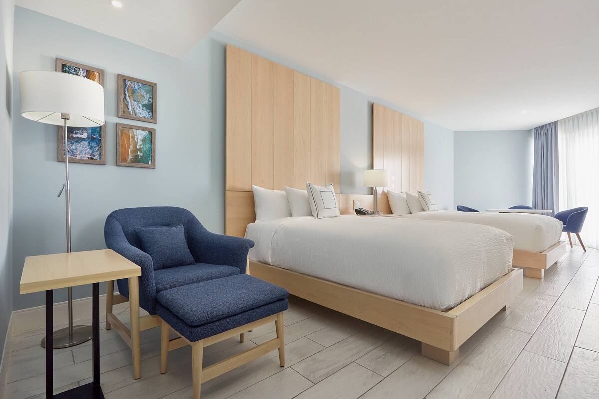 Una habitación de hotel moderna con una cama grande, un sillón azul, una mesita pequeña y obras de arte en la pared.
