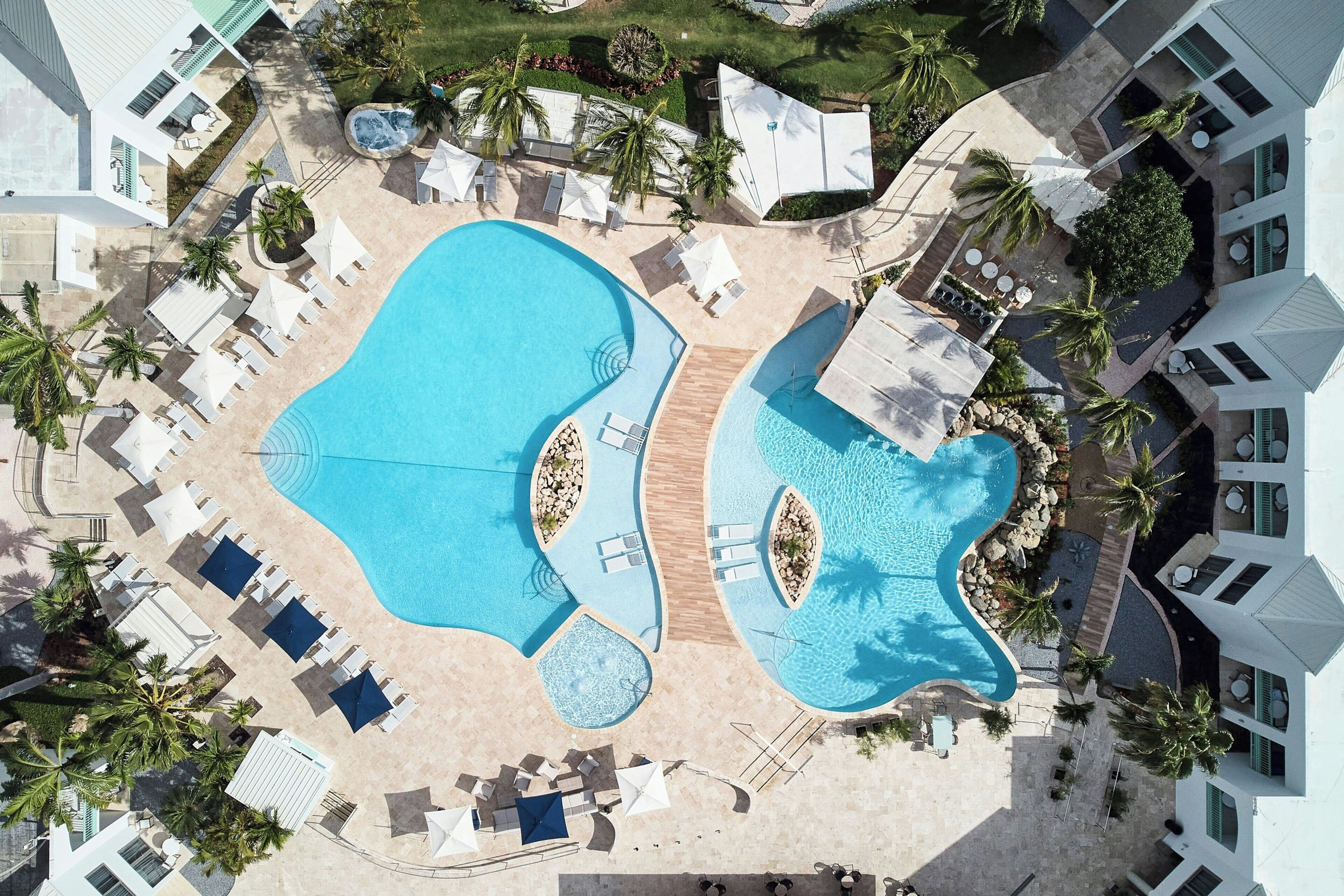 Oversigtsbillede af et resort med to store swimmingpools, liggestole og palmer.