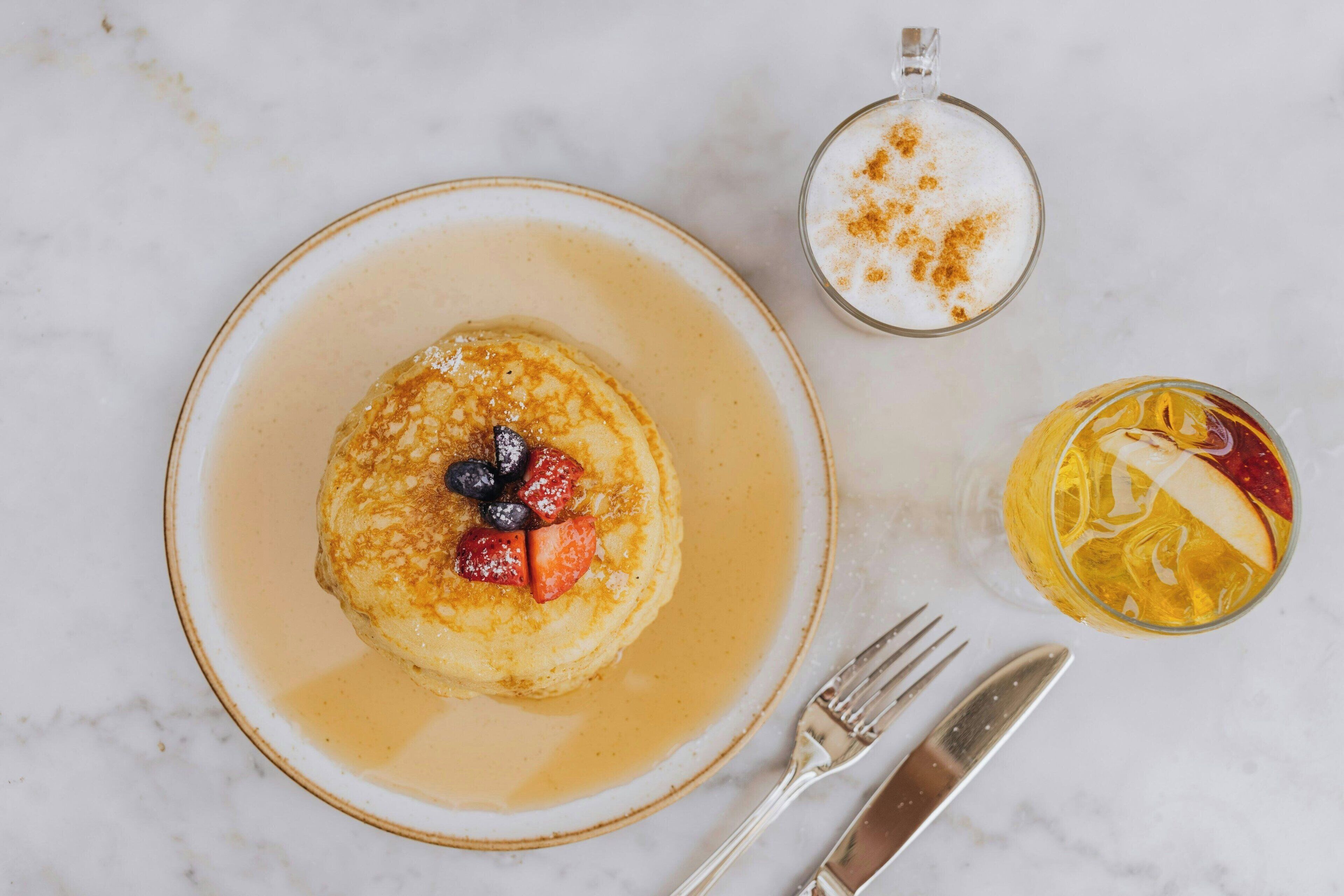 Une assiette de petit-déjeuner avec des crêpes, des myrtilles et du sirop, une tasse de café et un verre de jus d’orange garni de tranches de fruits.