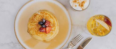 Une assiette de petit-déjeuner avec des crêpes, des myrtilles et du sirop, une tasse de café et un verre de jus d’orange garni de tranches de fruits.