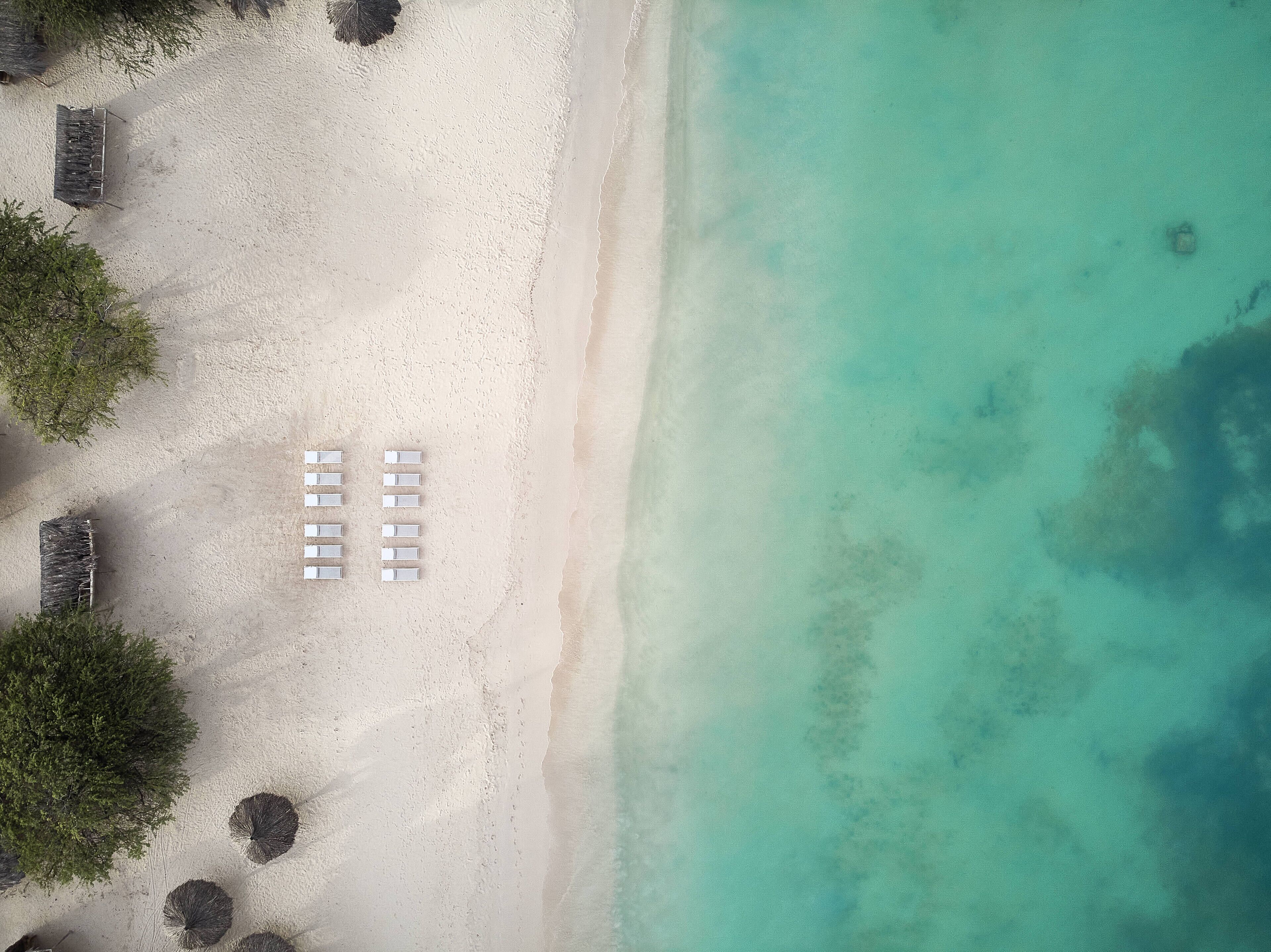 Vista aerea di una spiaggia con sabbia bianca, acque turchesi cristalline e una fila di sedie a sdraio bianche.