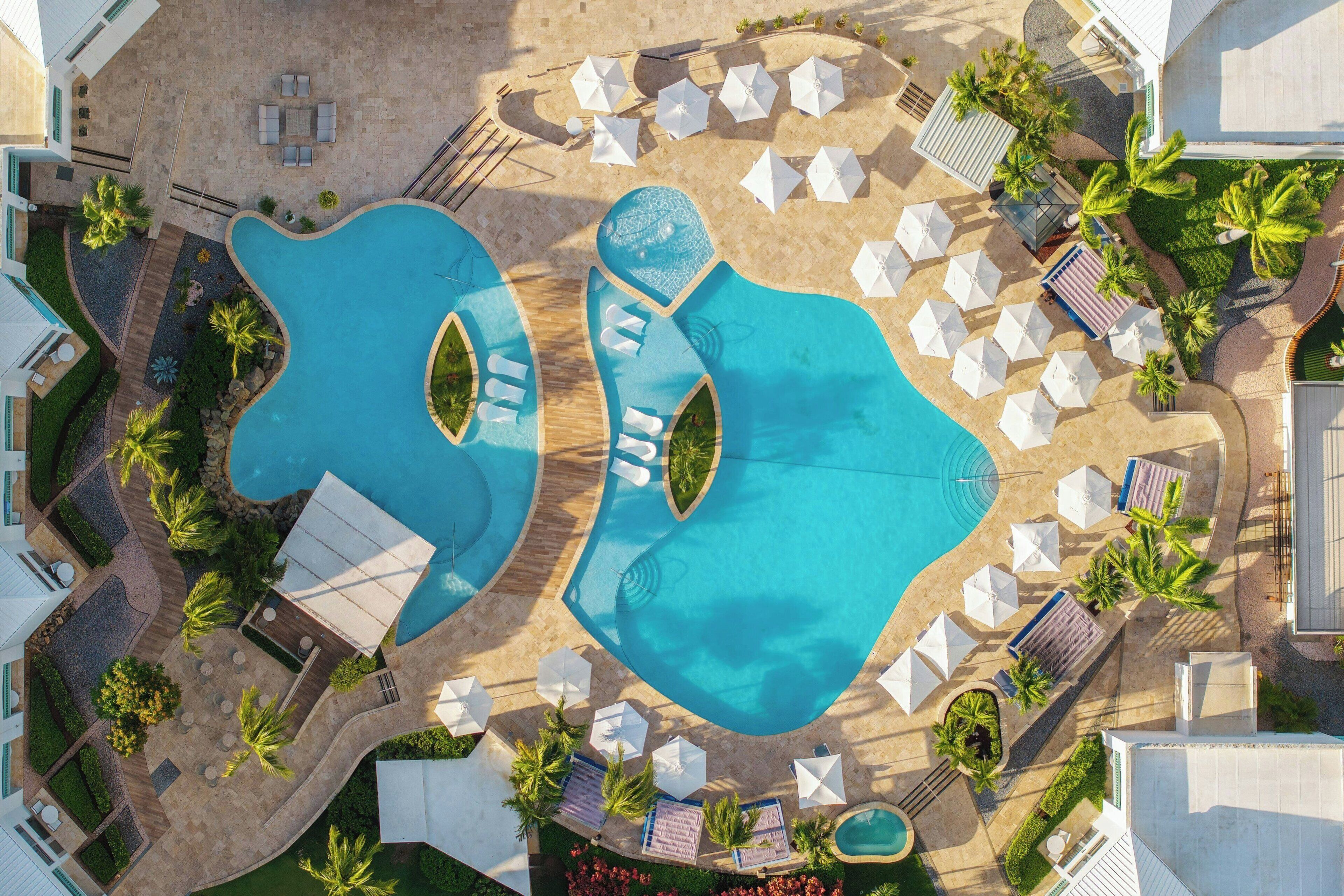 Vista aérea de un complejo turístico con dos piscinas grandes, tumbonas, sombrillas y palmeras.