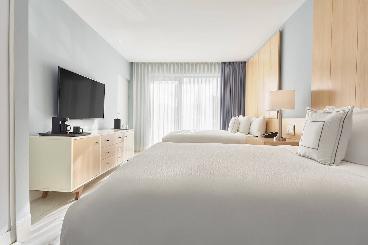 A hotel room with a large bed, a TV mounted on the wall, a bedside table with a lamp, and a window with sheer curtains.