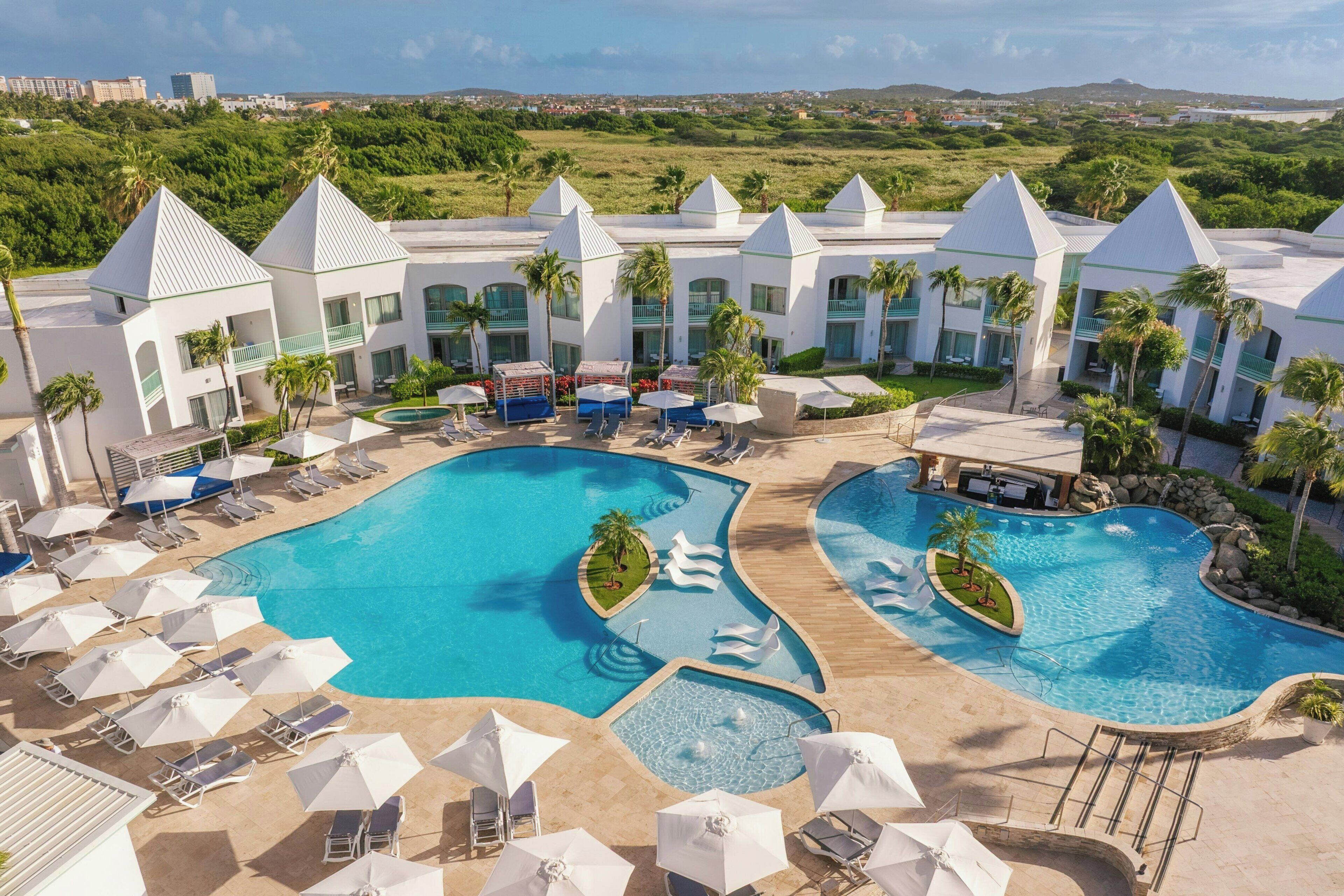 A resort with a large swimming pool, sun loungers, umbrellas, and a view of the surrounding landscape.