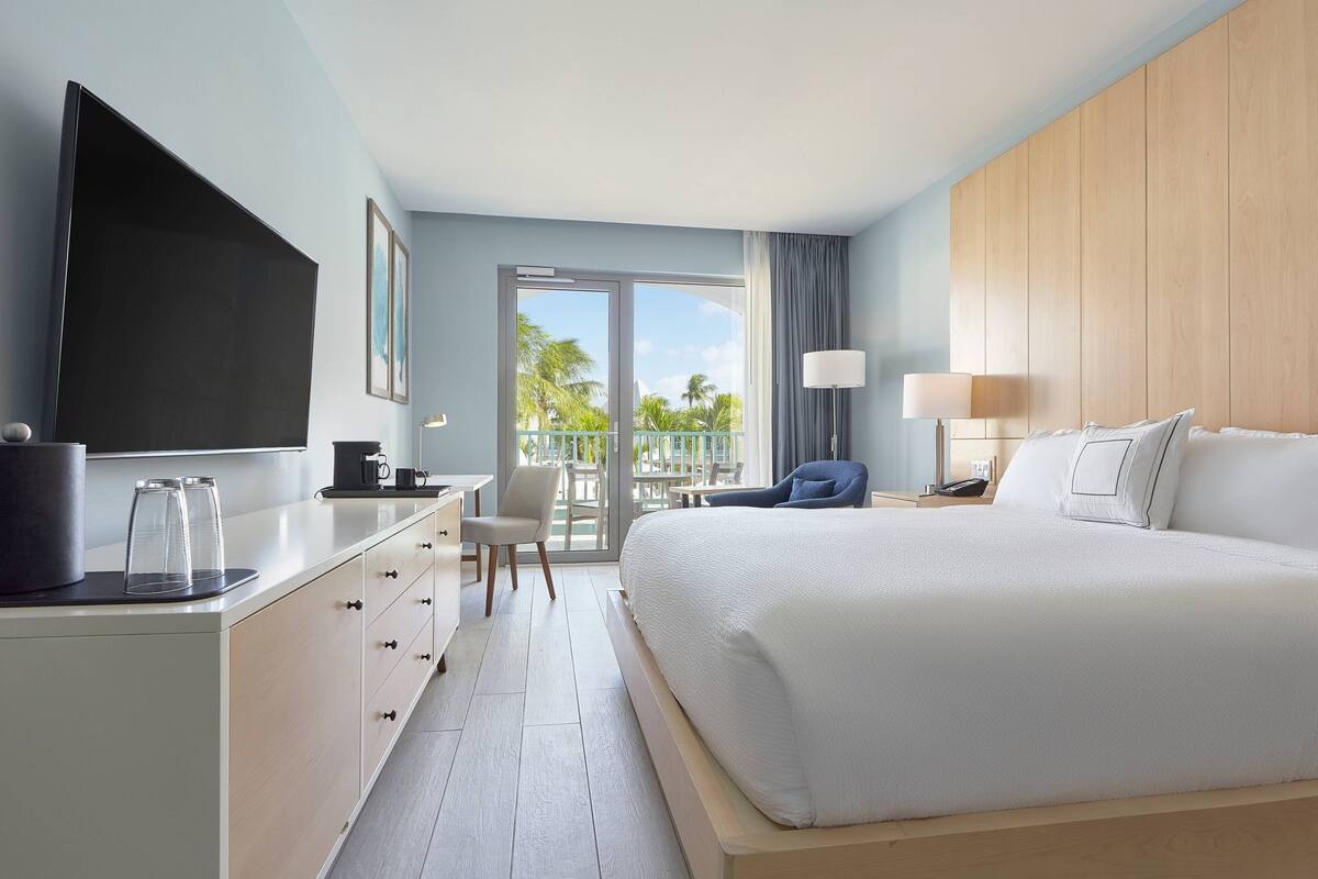 A modern hotel room with a large bed, a desk with a flat-screen TV, a chair, and a balcony with a view of palm trees.