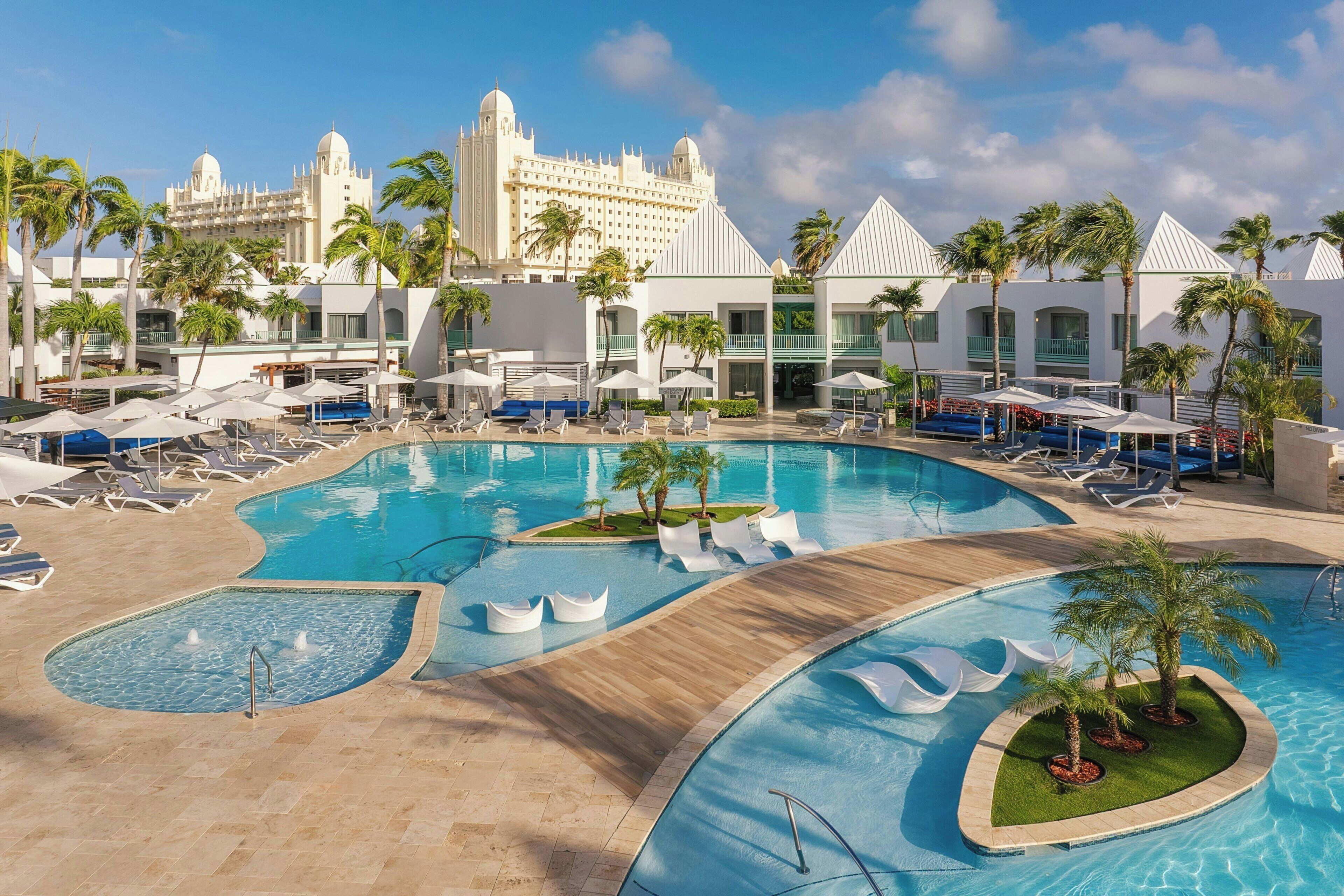 Un resort con varias piscinas, palmeras y un edificio grande al fondo.