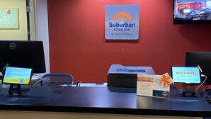 A reception desk with a sign for Suburban Studios by Choice Hotels, a computer, a printer, and a monitor displaying a welcome message.