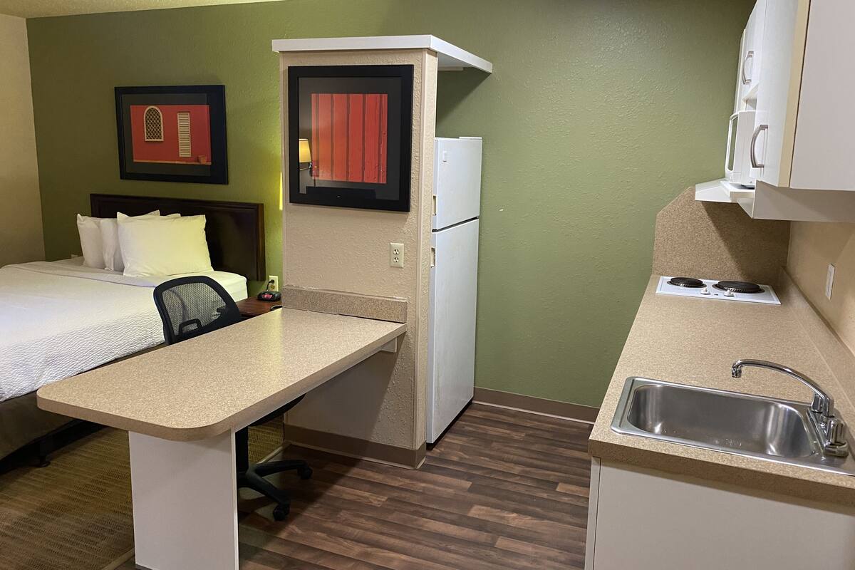 Une chambre d’hôtel de taille réduite, comprenant un lit, un petit bureau, une kitchenette et une seule chaise.