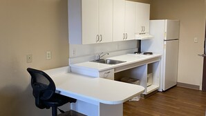 A compact kitchen with white cabinets, a sink, a refrigerator, and a black office chair.