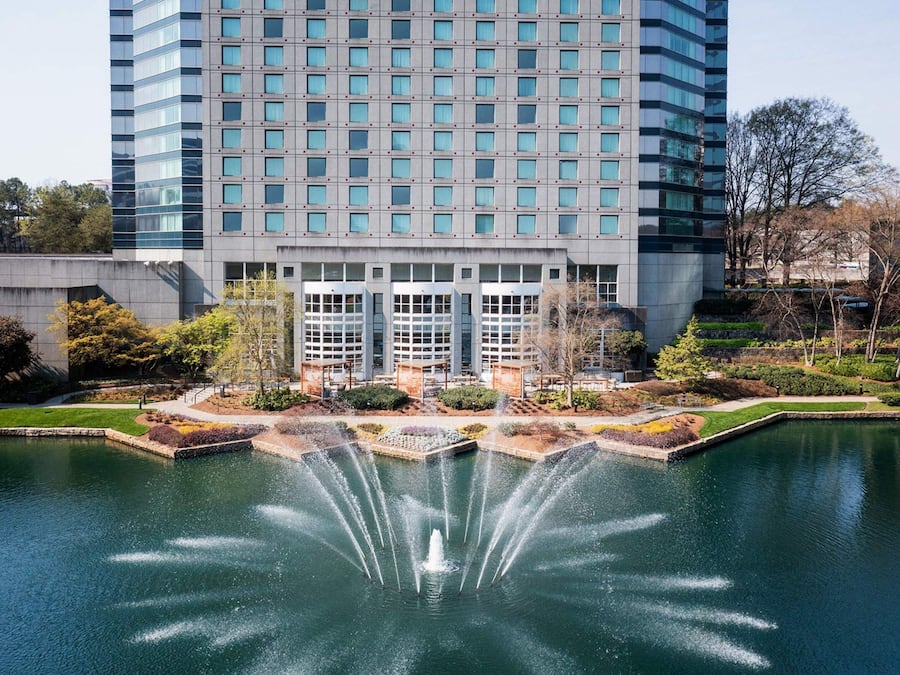 A modern hotel with a large water fountain in the courtyard, surrounded by greenery and a reflective pool.
