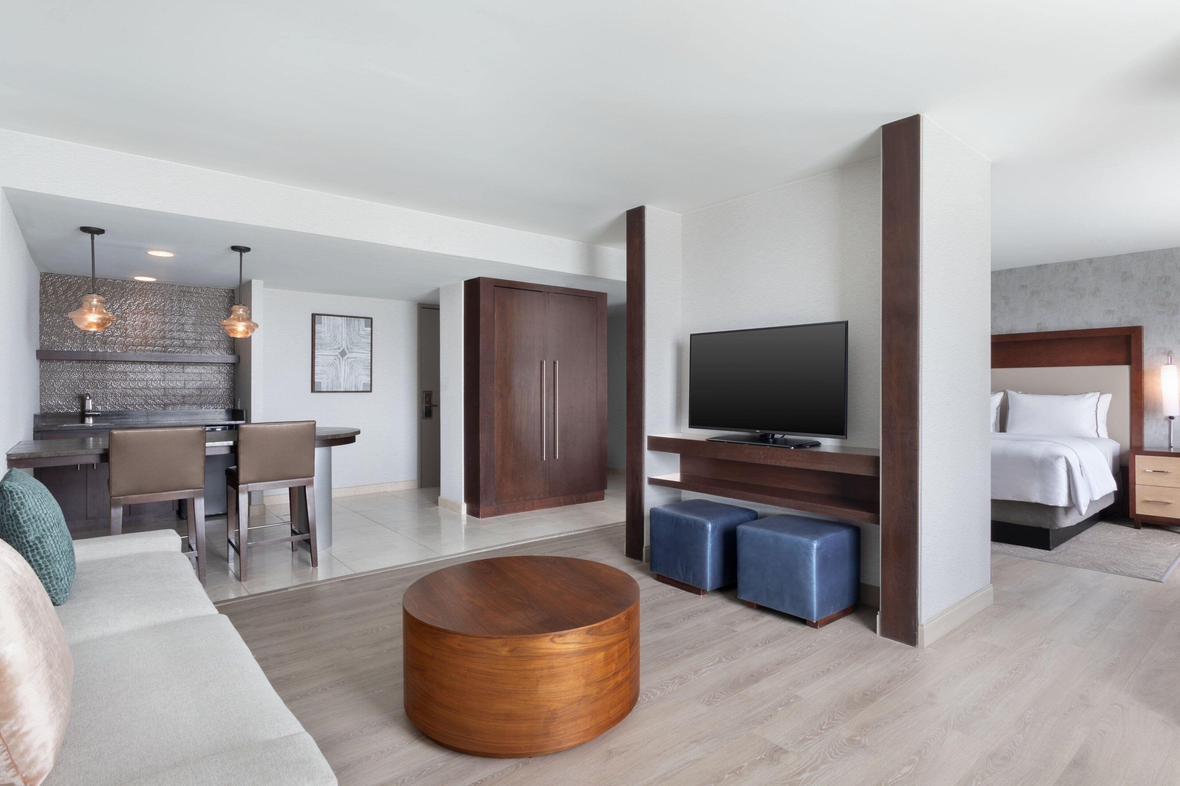 A modern hotel room with a kitchen area, a TV, a sofa, and a round wooden coffee table.