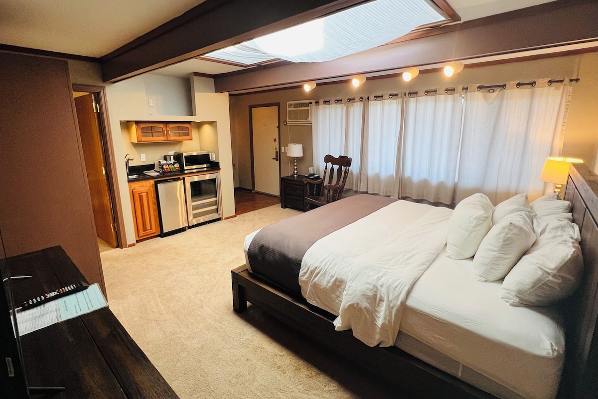 A hotel room with a large bed, a desk with a computer, a kitchenette, and a bathroom.