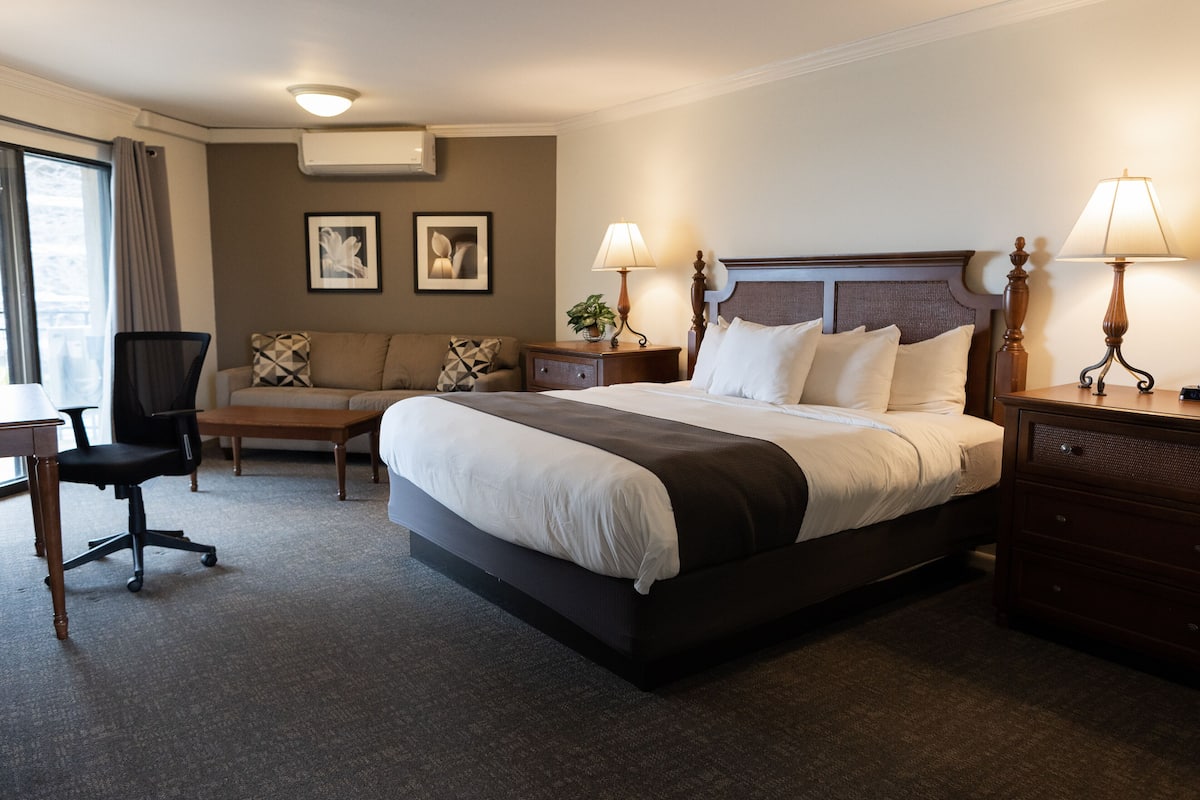 A hotel room with a large bed, two bedside tables with lamps, a desk, a chair, a sofa, and two framed pictures on the wall.