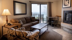 A living room with a sofa, a coffee table, a patterned armchair, a lamp, a phone, a framed mirror, a fireplace, and a balcony with a view.